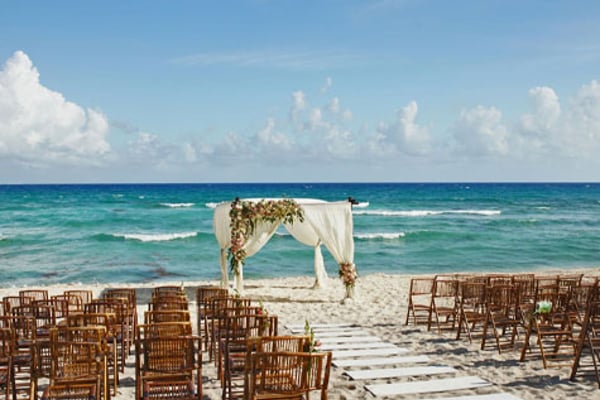 Beach wedding ceremony setup with wooden chairs facing a decorated arch on sand, turquoise ocean and white clouds beyond