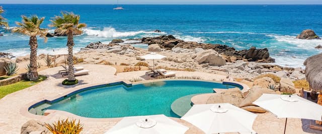 Villa de lujo con piscina infinita, tumbonas y terraza iluminada con vistas a una bahía costera al anochecer