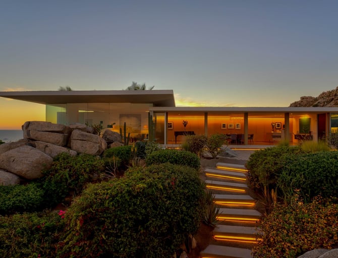 Casa moderna de una planta con paredes de cristal iluminada al anochecer, rodeada de jardines desérticos, grandes rocas y un sendero iluminado con vistas al mar al atardecer.